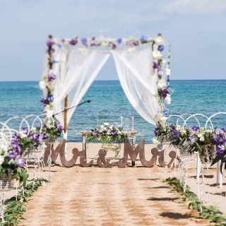 Nice view of a decorated path for the wedding ceremony on a beach. Violet flowers. Sea, sky and Mr Mrs plate on background.